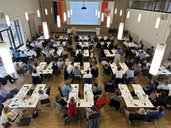 Blick auf den gesamten Mehrzwecksaal mit Teilnehmern an Tischen und der Bühne mit Leinwand